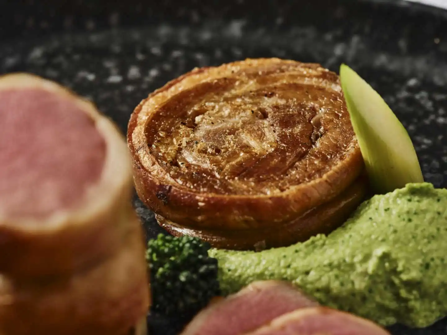 Crispy rolled pork belly with pea puree, charred broccoli and apple on a dark plate at a fine dining catered event in Bournemouth